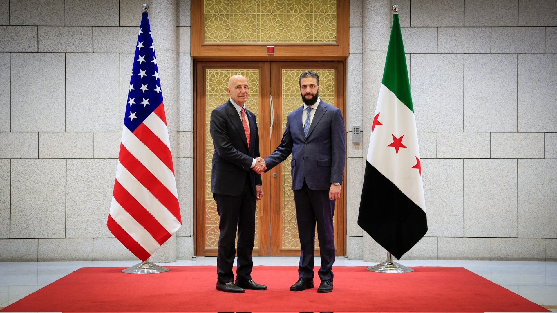 US Special Envoy Tom Barrack shakes hands with Syrian President Ahmad al-Sharaa at the People's Palace in Damascus, May 2025, with US and Syrian flags in the background