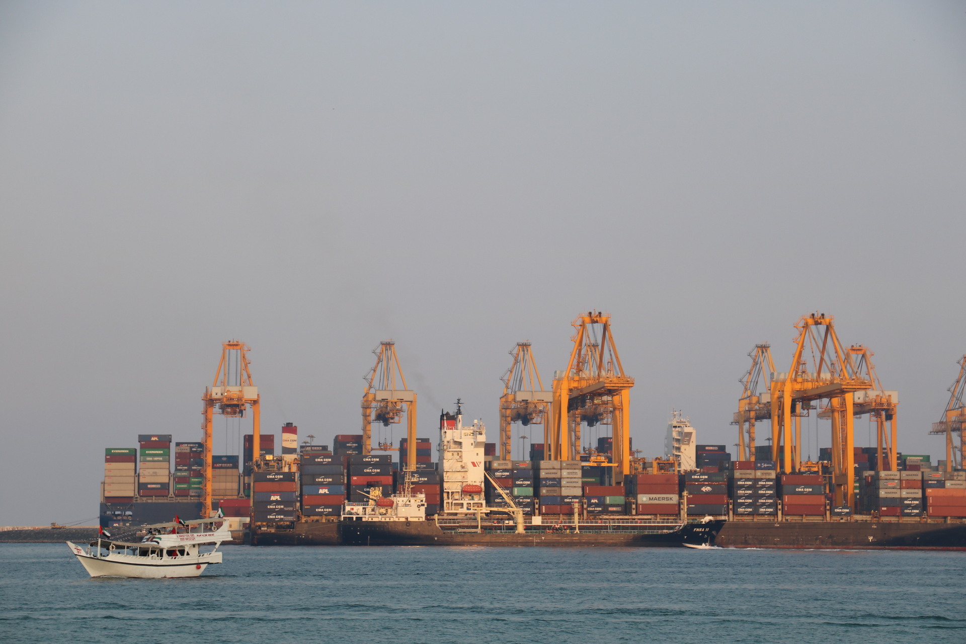 Fujairah Port container terminal showing shipping cranes and stacked containers, with small UAE-flagged dhow in foreground