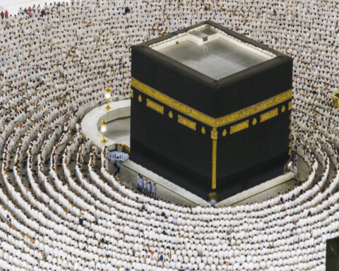 Pilgrims in white ihram garments in concentric rows of prayer surrounding the Kaaba at the Grand Mosque in Mecca, 2026. The 1.8 million pilgrims attending Hajj 2026 pass through the gap between Washington’s OSAC Level 3 advisory and Riyadh’s Patriot deployment photographs — a gap the Saudi Ministry of Defence has expressed in imagery rather than interceptor counts.