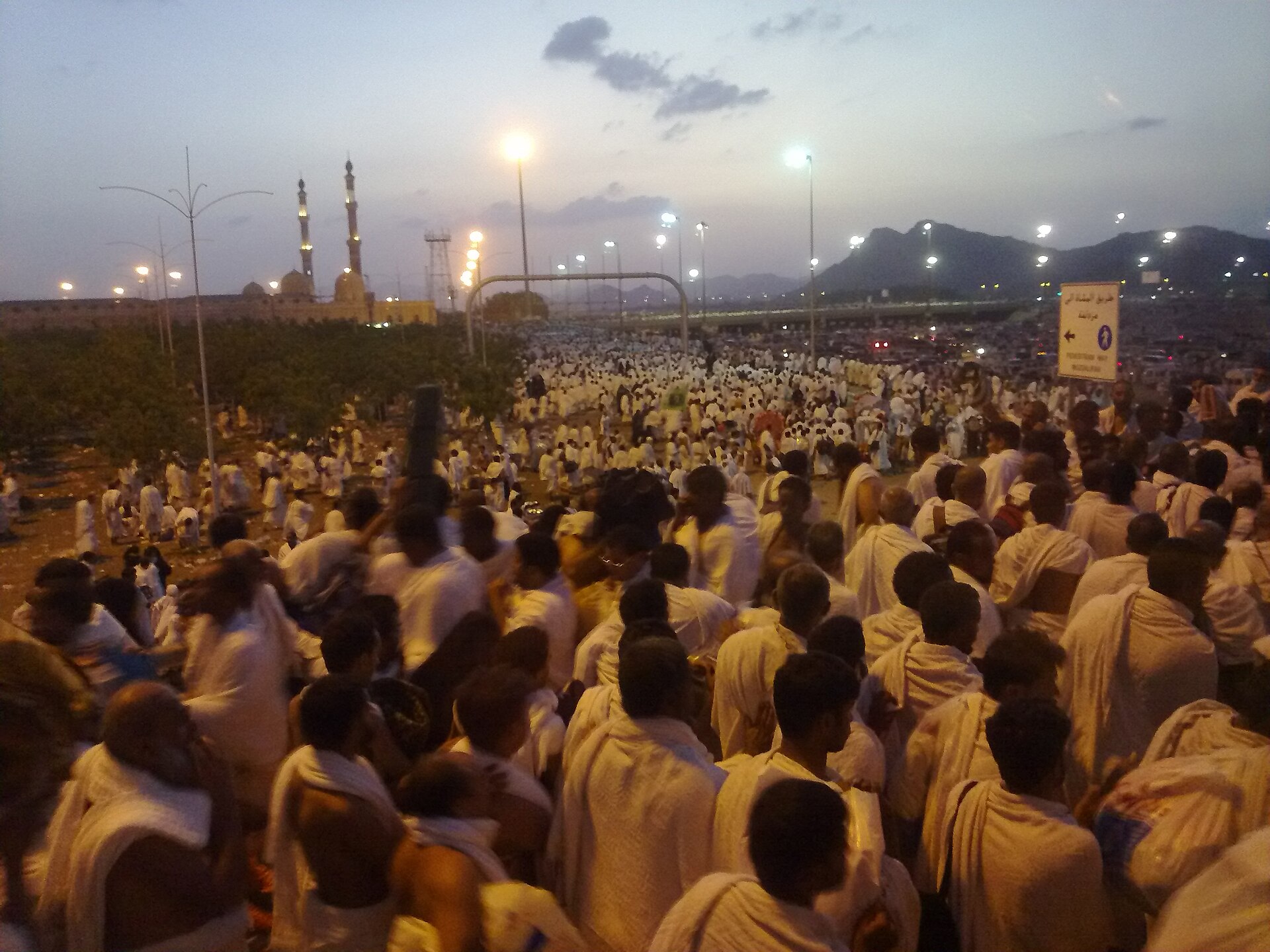 Hajj pilgrims in white ihram garments leaving Arafat plain for Muzdalifah at sunset, with mosque and Jabal al-Rahmah visible in background