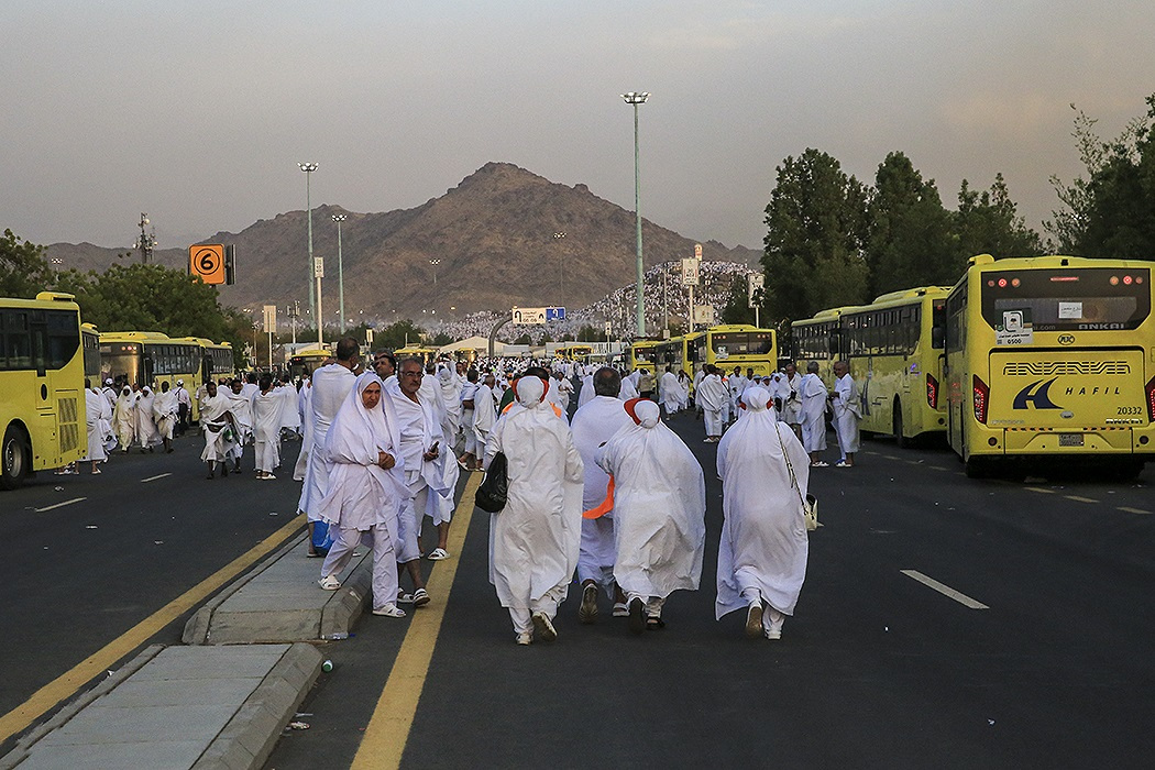 Hajj pilgrims in white ihram walking from Arafat plain toward transport buses, with Jabal al-Rahmah (Mount of Mercy) visible in background, August 2018