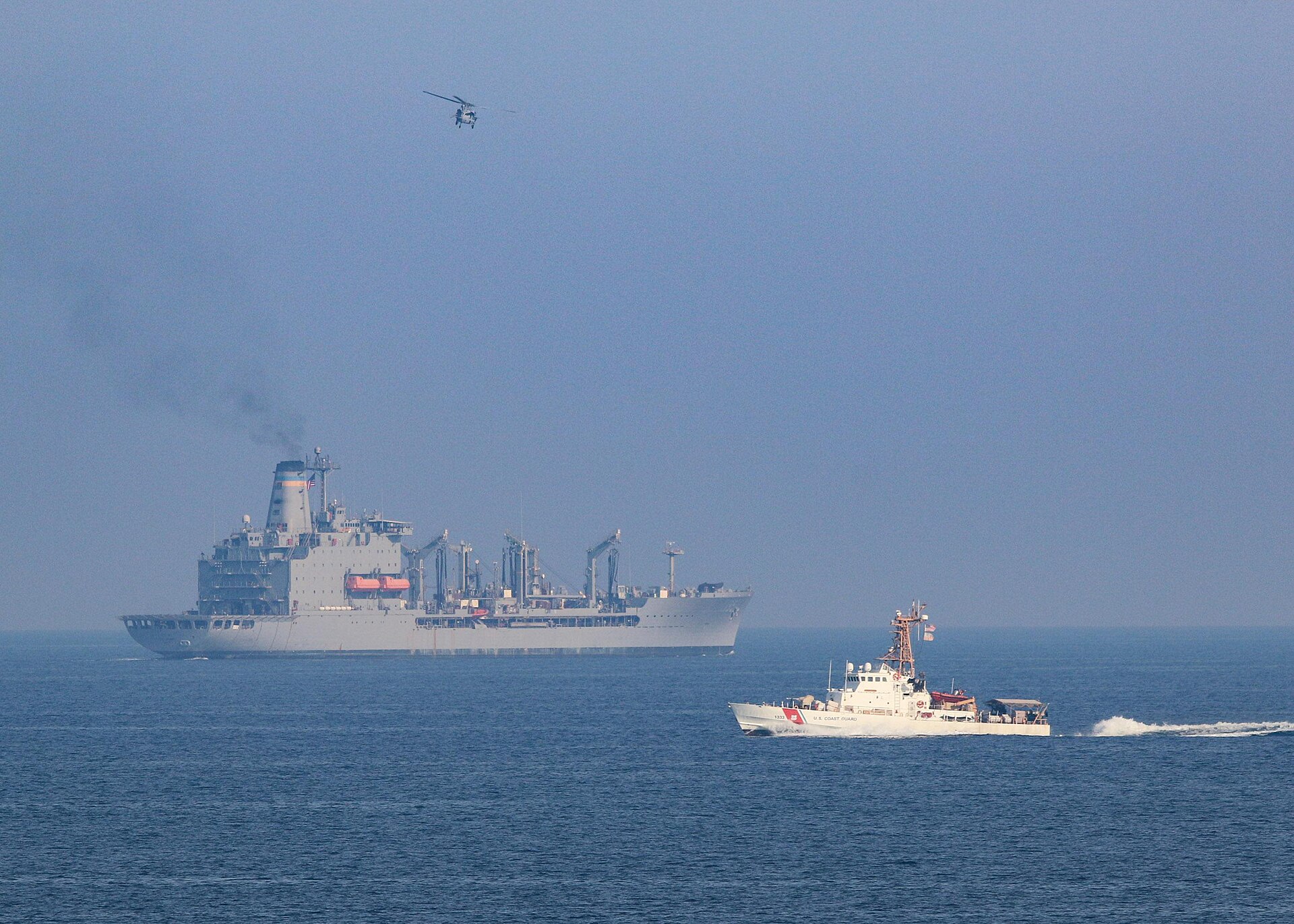 USCGC Adak and USS Winston S. Churchill DDG-81 operating in the Persian Gulf in September 2020, with a Navy helicopter overhead — part of the US Fifth Fleet presence that has escorted more than 60 commercial vessels since the blockade began in April 2026