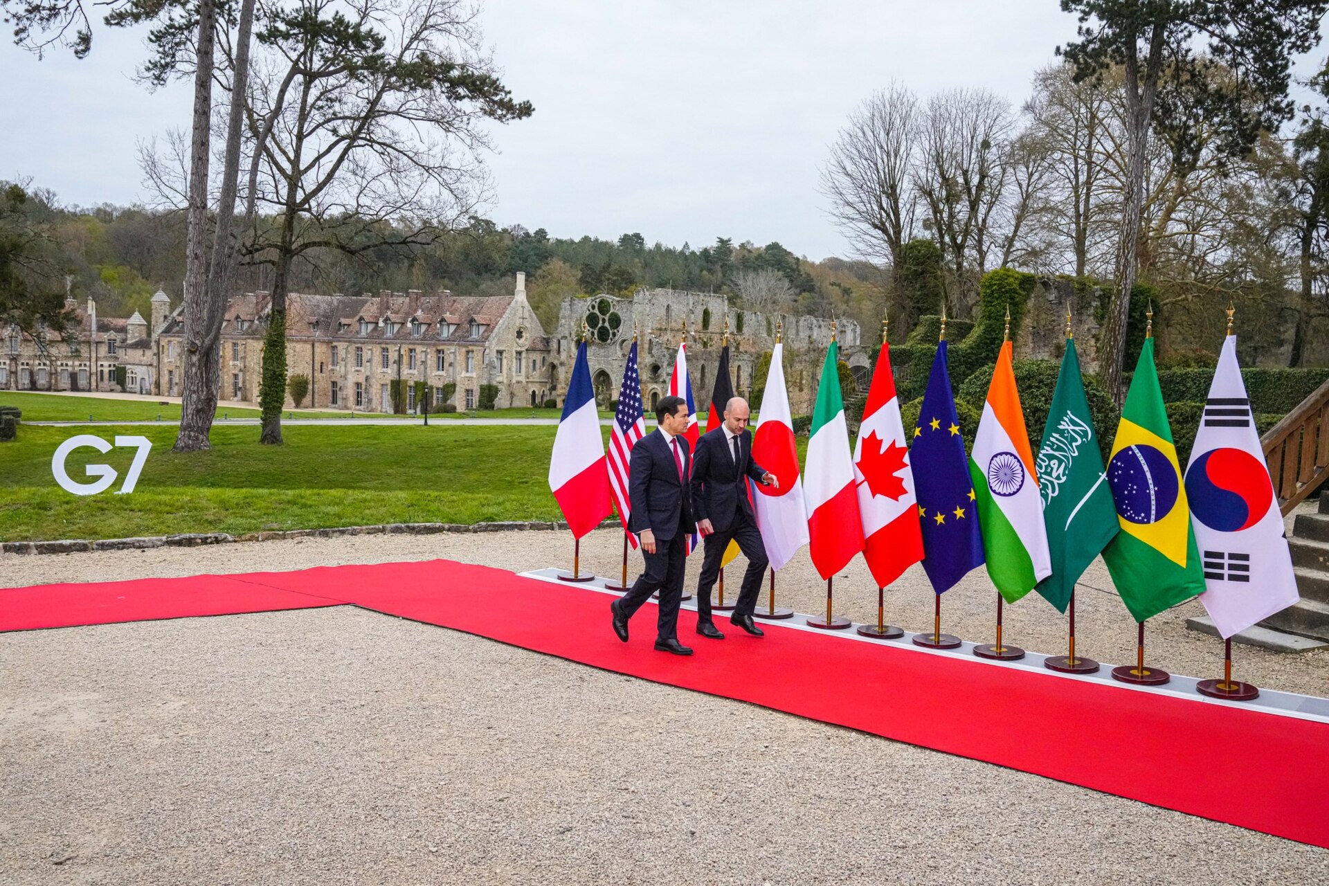 Secretary Rubio greeted by French Foreign Minister Barrot at G7 Foreign Ministers meeting Abbaye des Vaux-de-Cernay March 2026, with Saudi Arabian flag visible among G7-plus delegation banners