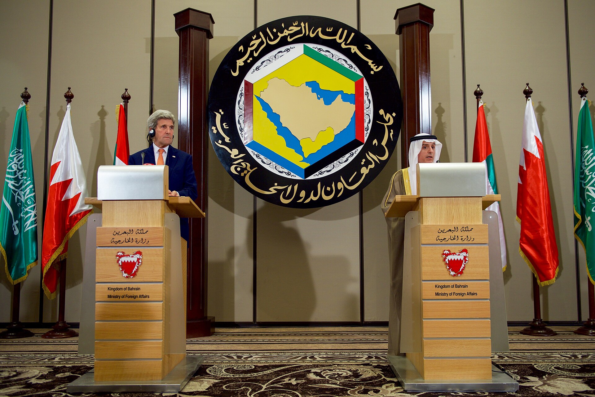 US Secretary of State Kerry and Saudi FM Adel al-Jubeir at dual podiums beneath the GCC emblem at Bahrain's Foreign Ministry in Manama, after a Gulf Cooperation Council ministerial meeting — the same venue framework used for the June 25, 2026 GCC 167th Ministerial Council that produced the council's first collective defense language