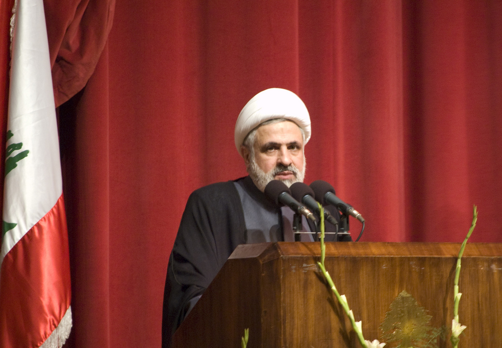 Naim Qassem, then Hezbollah Deputy Secretary-General, speaks at a podium with the Lebanese cedar flag behind him — he made his rejection of the June 4 ceasefire explicit and public, using the word surrender