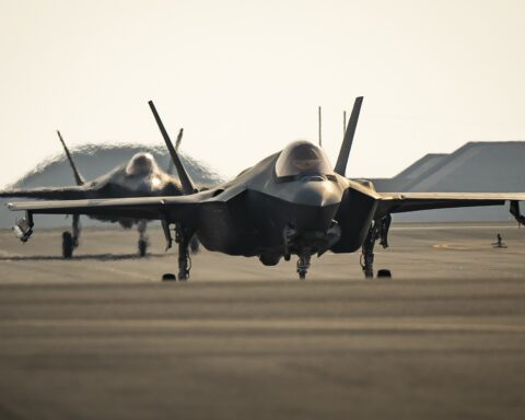 Two F-35A Lightning II stealth fighters taxi on the runway at Al Udeid Air Base, Qatar, with hardened aircraft shelters visible in the background