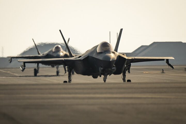 Two F-35A Lightning II stealth fighters taxi on the runway at Al Udeid Air Base, Qatar, with hardened aircraft shelters visible in the background