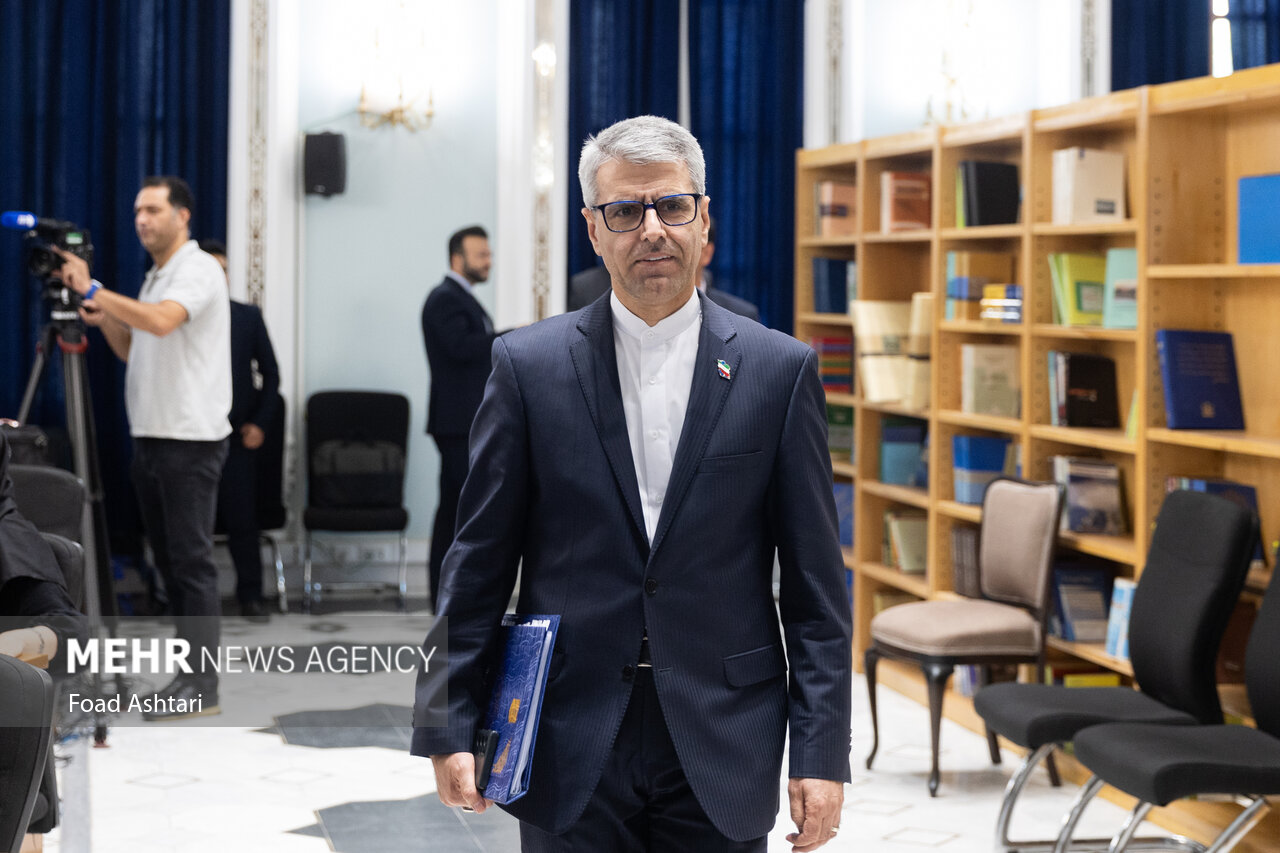 Iranian Foreign Ministry spokesperson Esmaeil Baghaei arrives at the Tehran MFA briefing room carrying a dossier, with Iranian flags and media equipment visible in the background