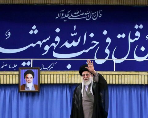 Ayatollah Ali Khamenei addresses a public gathering at Imam Khomeini Hosseinieh, waving from the podium beneath a Quranic inscription. Khamenei holds sole constitutional authority over war and peace under Article 110 of the Iranian constitution.