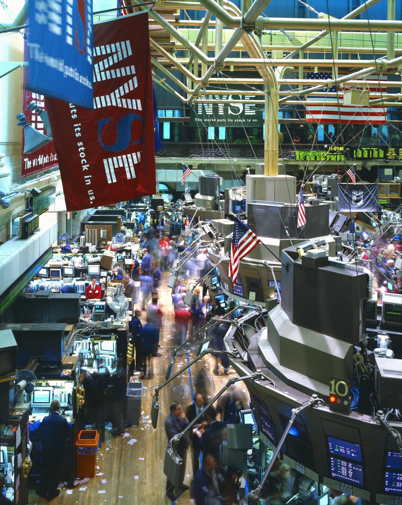 New York Stock Exchange trading floor with brokers at workstations, financial screens displaying market data, American flags overhead
