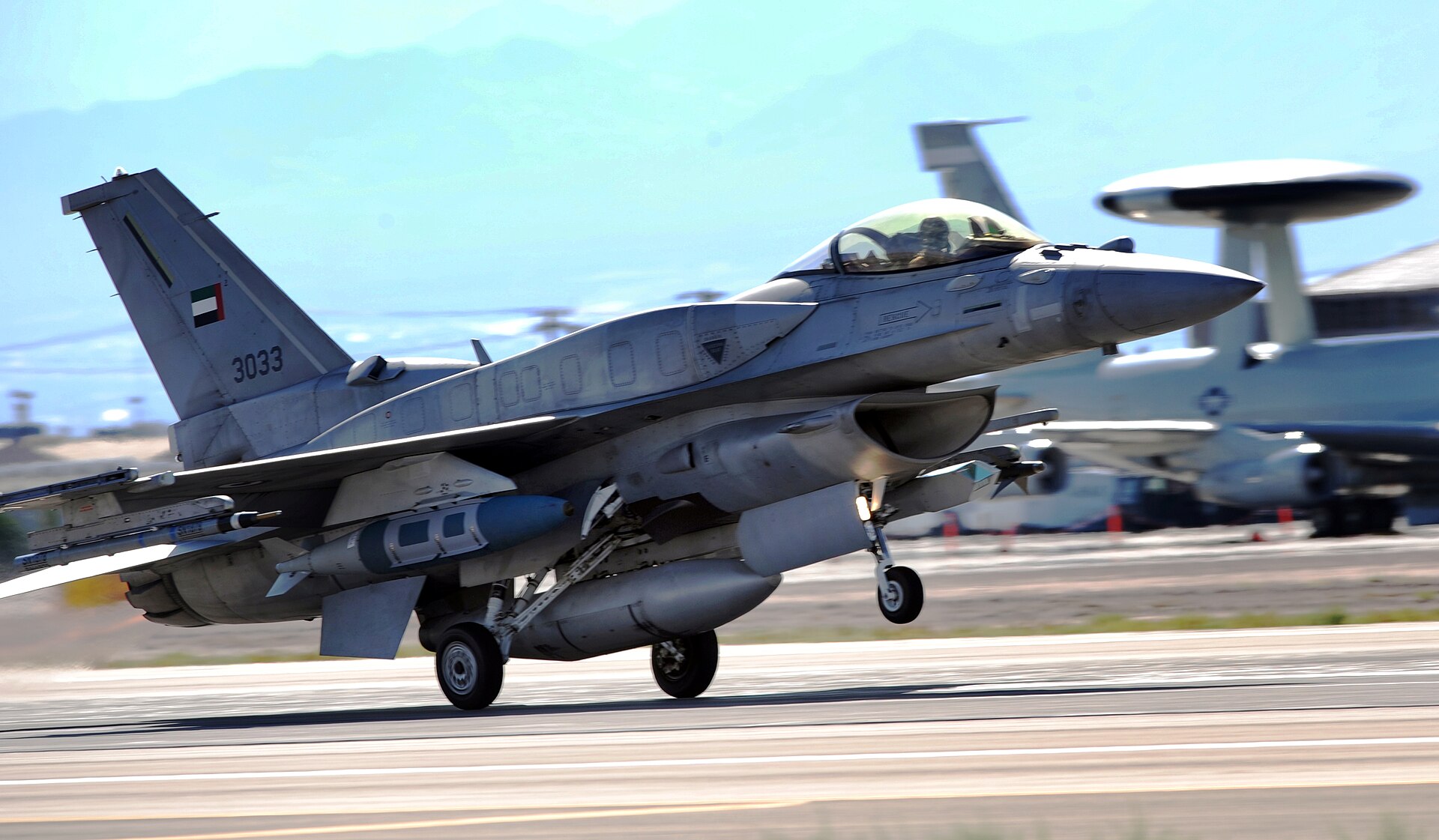 UAE Air Force F-16E Block 60 fighter jet with UAE flag on tail, an AWACS early warning aircraft visible in background — the UAE operates its full air-defense network from Al Dhafra Air Base near Abu Dhabi