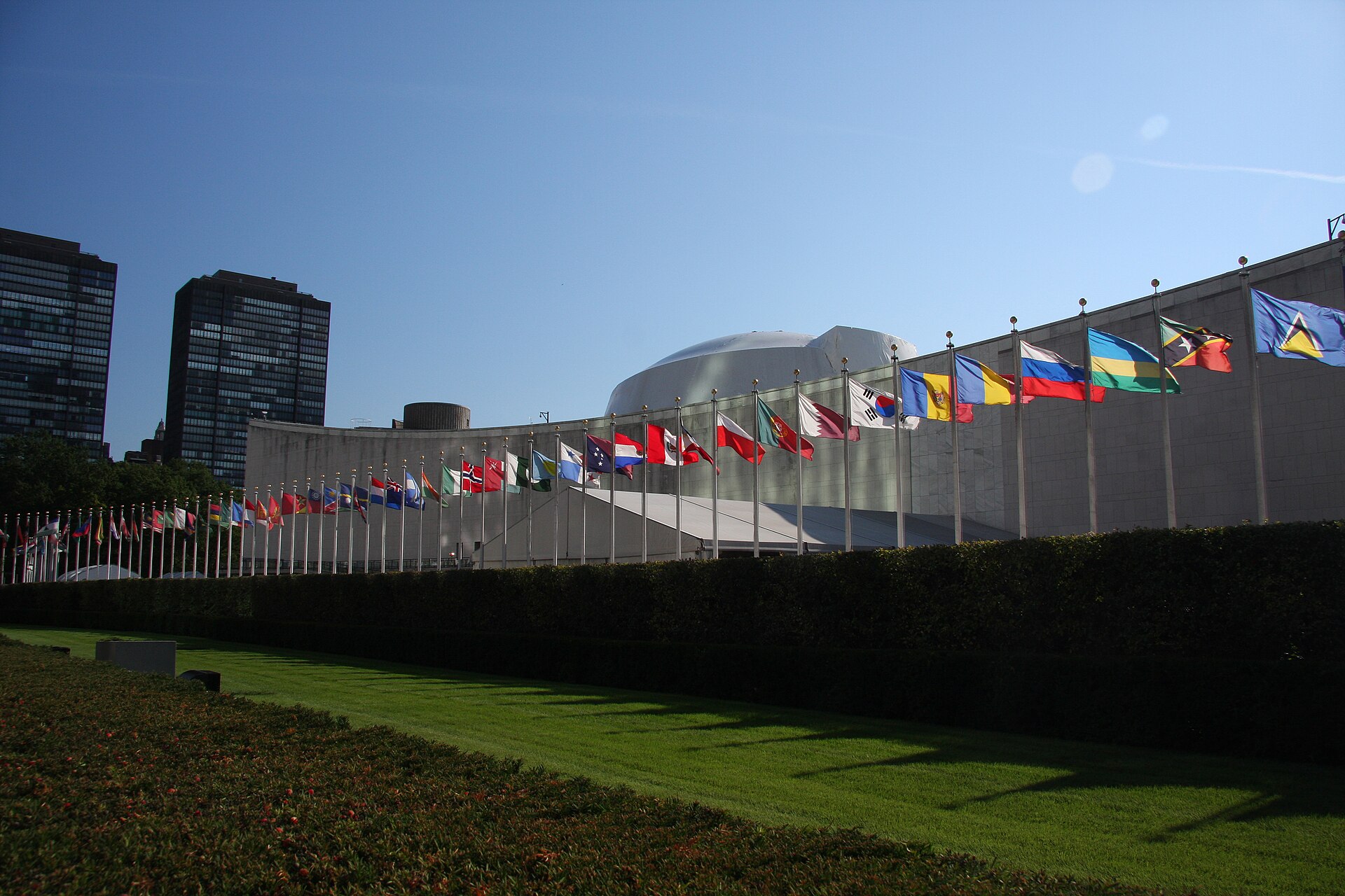 Flags of United Nations member states flying outside the UN General Assembly building in New York, with the distinctive dome of the Security Council chamber visible in the background