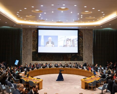The UN Security Council chamber during a full session, viewed from above, showing the horseshoe table with all member state delegates seated and a projection screen at the far end