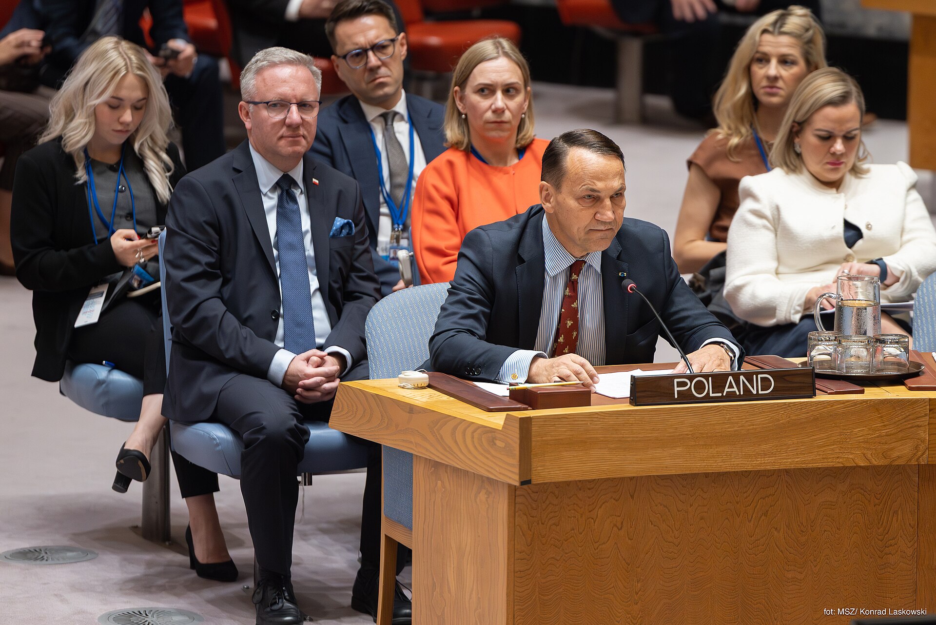 A delegate addresses the UN Security Council during an emergency session, with the POLAND nameplate visible at the horseshoe table and other member states' representatives seated behind