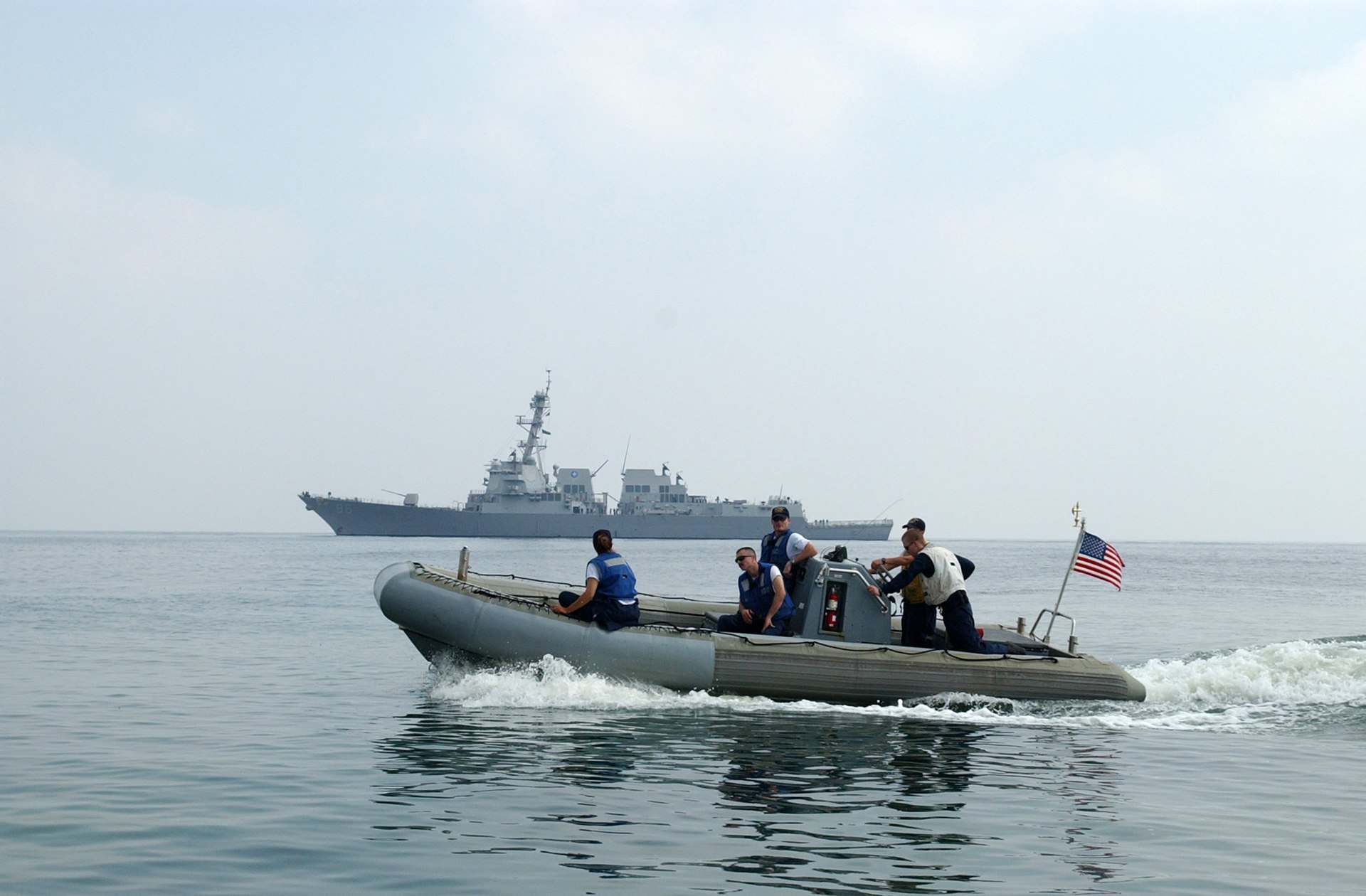 US Navy sailors in a rigid-hull inflatable boat patrol waters near Yemen coast with guided-missile destroyer USS Bainbridge in background during search and rescue operations