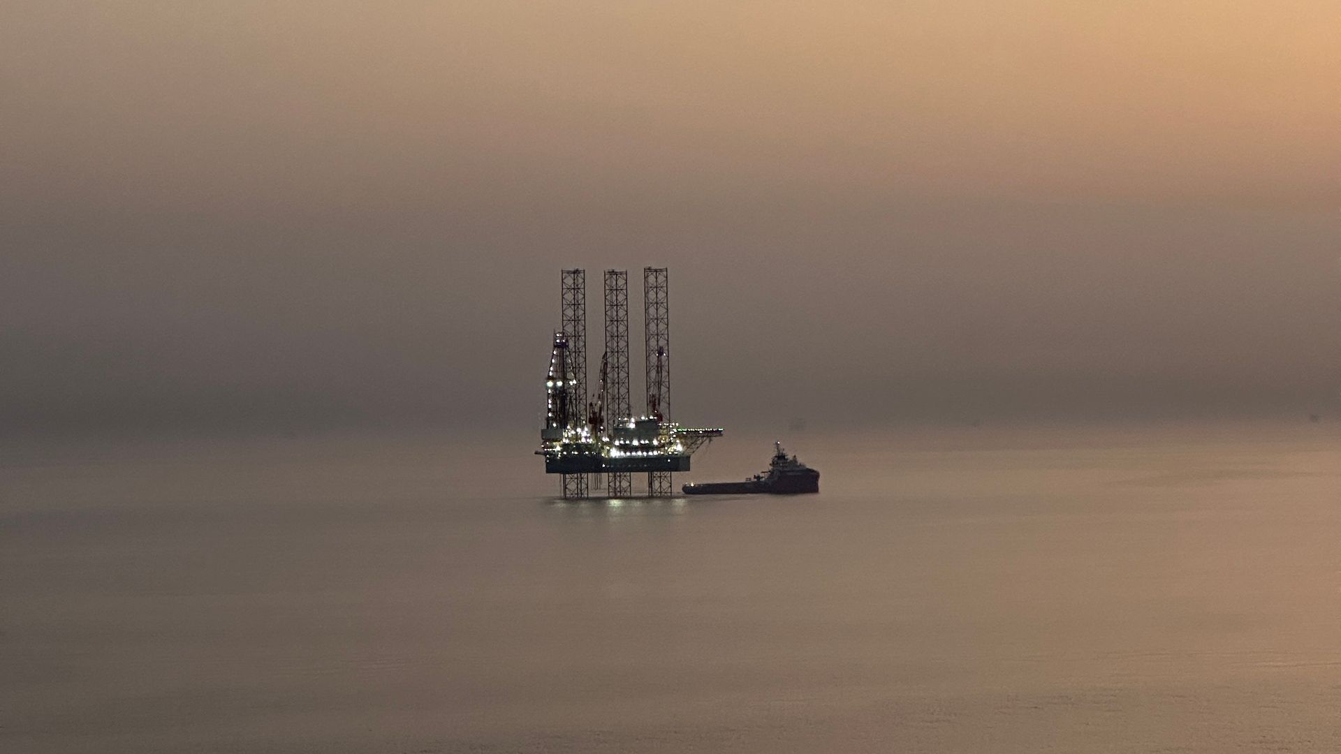 A Saudi Aramco offshore oil rig lit against a hazy Gulf sunset, with a supply vessel moored alongside — the Eastern Province holds the world's largest proven oil reserves