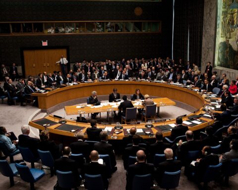 UN Security Council members seated at the horseshoe table during a formal session, with country placards and observers filling the chamber